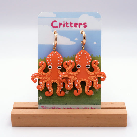 Orange octopus-shaped earrings on a 'Critters' branded card with a wooden stand.