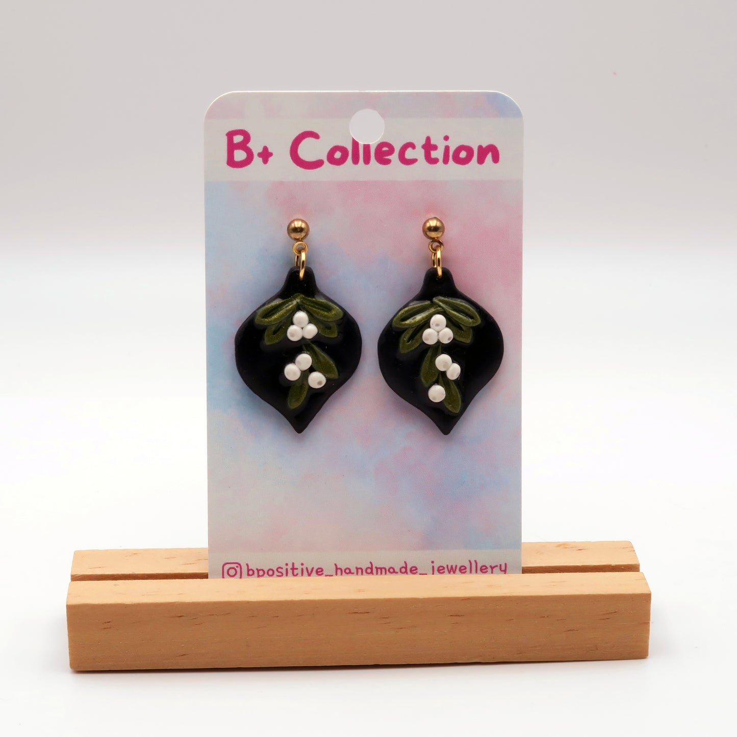Black bauble shaped mistletoe polymer clay earrings,  on a 'B+ Collection' card, displayed on a wooden stand.